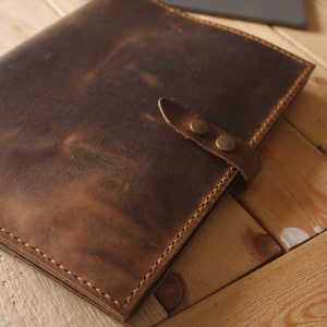 May include: A brown leather folder with a strap and two brass snaps. The folder is closed and lying on a wooden surface.
