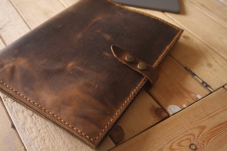 May include: A brown leather folder with a strap and two brass snaps. The folder is closed and lying on a wooden surface.
