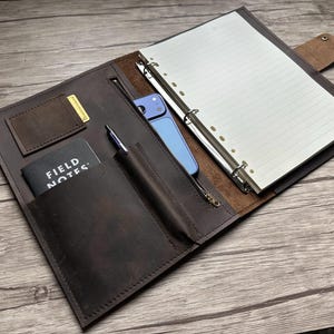 May include: A dark brown leather organizer folio featuring a six-ring binder with lined paper, a zippered pocket containing a blue smartphone, a pen loop with a pen, a small pocket with a card, and a Field Notes notebook in a front sleeve.