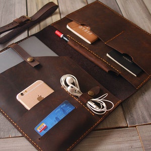 May include: A brown leather portfolio with multiple pockets and compartments. The portfolio is open and contains a tablet, phone, credit card, and headphones.