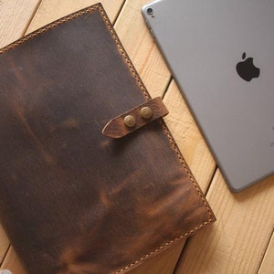 May include: A brown leather notebook with a strap and metal clasps. The notebook is lying on a wooden surface next to an iPad.