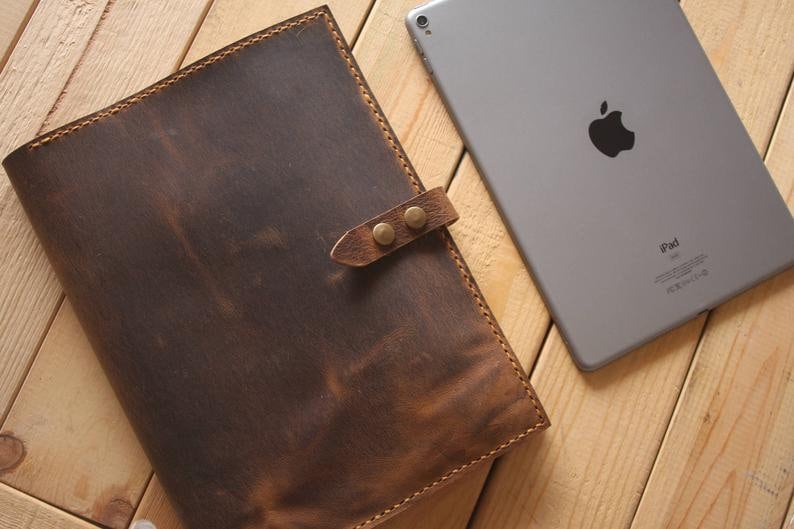 May include: A brown leather notebook with a strap and metal clasps. The notebook is lying on a wooden surface next to an iPad.