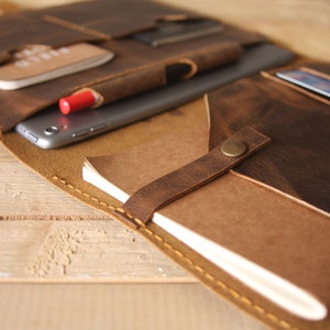 May include: A brown leather portfolio with multiple pockets and compartments. The portfolio is open and shows a tablet, a notebook, a pen, and a credit card.