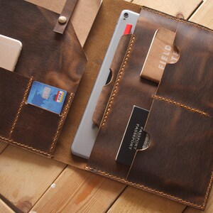 May include: A brown leather portfolio with multiple pockets and a strap. The portfolio is open to reveal a tablet, a passport, a credit card, a phone, and a notebook.