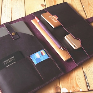 Könnte beinhalten: Ein dunkelviolettes Lederportfolio mit mehreren Taschen und Fächern. Das Portfolio ist geöffnet und enthält ein Tablet, ein Notizbuch, einen Stift, eine Kreditkarte, einen Reisepass und ein Telefon.