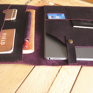 Op de afbeelding: Een paarse leren portfolio met meerdere zakken en vakken. De portfolio is open en bevat een tablet, een notitieboekje, een pen, een telefoon en een paspoort.