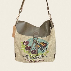 May include: A beige canvas tote bag with a brown leather strap and trim. The bag features a vintage-style print of a bee and flowers with the text "SELECTED FOR PURITY - GERMINATION ADAPTABILITY Plymouth, Mich. 25 lbs."