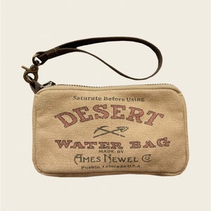 May include: A tan canvas water bag with a brown strap and zipper closure. The bag has the text "Saturate Before Using" and "DESERT WATER BAG" printed in pink. The text "MADE BY AMES NEWEL C" and "Pueblo, Colorado U.S.A." is printed in black.