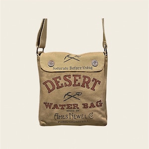 May include: A tan canvas water bag with the text "Desert Water Bag" in red and brown lettering. The bag has a flap with the text "Saturate Before Using" and a small pocket with a button closure. The bag is made by Ames Newell Co. in Pueblo, Colorado, USA.
