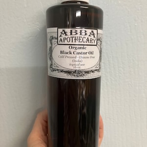 Può includere: Una bottiglia marrone scuro di ABBA Apothecary Organic Black Castor Oil. L'etichetta include il testo "Cold Pressed - Hexane Free (India) topical use 16 oz." La bottiglia ha un tappo bianco ed è tenuta da una mano.