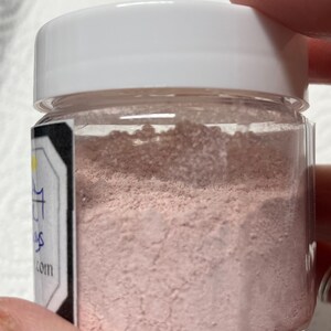 Pu&ograve; includere: Un piccolo barattolo di vetro trasparente con un coperchio bianco riempito di polvere rosa chiaro. Il barattolo ha un'etichetta con il testo "Sugar & Spice" e un indirizzo di sito web.
