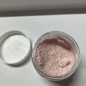 Pu&ograve; includere: Un piccolo barattolo trasparente pieno di polvere rosa chiaro. Il barattolo ha un coperchio bianco.
