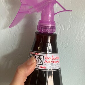 Può includere: Una bottiglia di vetro trasparente con un ugello spray viola riempito di un liquido scuro. La bottiglia ha un'etichetta bianca con il testo "Very Gentle Magnesium Oil" e "www.AbbaApothecary.com Daytona Beach, Florida".