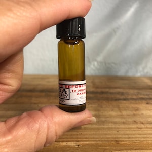 Può includere: Una piccola bottiglia di vetro trasparente con tappo nero, tenuta tra le dita. La bottiglia contiene un liquido color oro e ha un'etichetta bianca con testo nero. L'etichetta recita "BA SCARY" e "10 DROPS CARRIER".