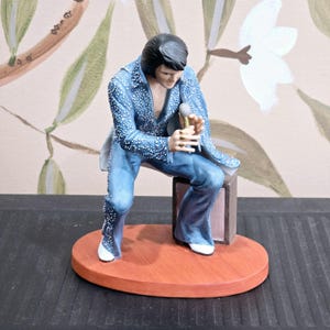 May include: A blue and silver figurine of Elvis Presley holding a microphone, sitting on a brown amplifier. The figurine is on a wooden base.