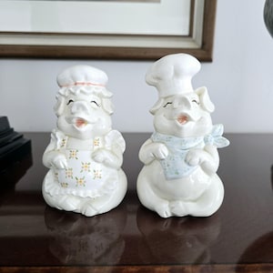 May include: A pair of white ceramic pig salt and pepper shakers. The pig on the left is wearing a white chef's hat and a floral apron. The pig on the right is wearing a white chef's hat and a blue bandana.