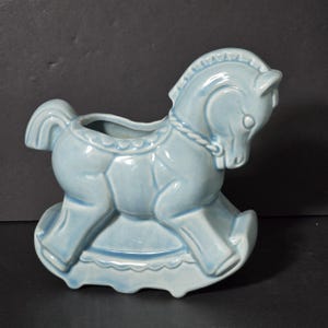 May include: A light blue ceramic rocking horse planter. The horse has a detailed mane and tail, and the base is shaped like a rocking horse.