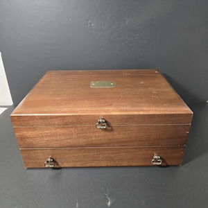 Może przedstawiać: Drewniana skrzynia dwupoziomowa z okuciami mosiężnymi. Górna część skrzyni ma mosiężną płytkę do grawerowania.