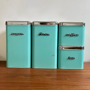 May include: A collection of four vintage aqua kitchen storage tins with silver lids. The tins are labelled "sugar", "flour", "coffee", and "tea" in a cursive script. The rectangular tins are displayed on a wooden surface.