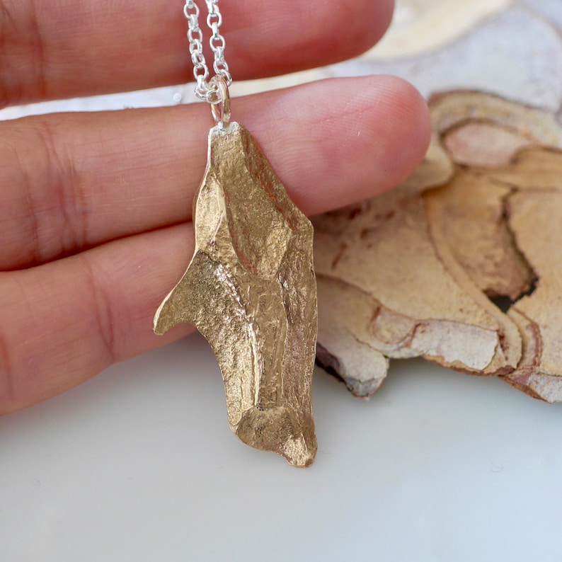 May include: A gold pendant necklace with a unique, abstract, textured design. The pendant is shaped like a jagged, asymmetrical rock or shard. The necklace is made of silver chain.
