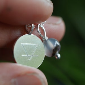 May include: Silver necklace with a round pendant featuring an engraved symbol and a grey agate bead.