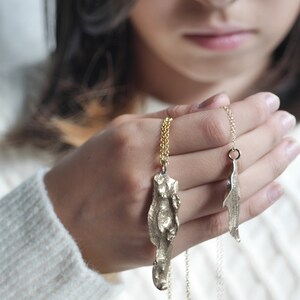 May include: Two gold necklaces with abstract pendant charms. The larger pendant is a textured, organic shape, while the smaller pendant is a more simple, elongated shape.