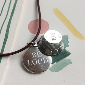 May include: A brown leather necklace with two silver charms. One charm is a round disc with the words "BE LOUD" engraved on it. The other charm is a silver guitar knob with the word "TONE" engraved on it.