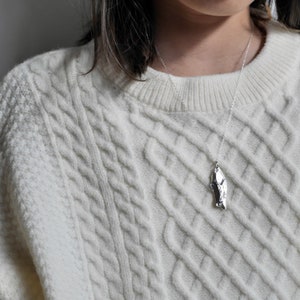 May include: A silver necklace with a unique, textured pendant shaped like a jagged rock. The necklace is worn by a person wearing a white cable knit sweater.
