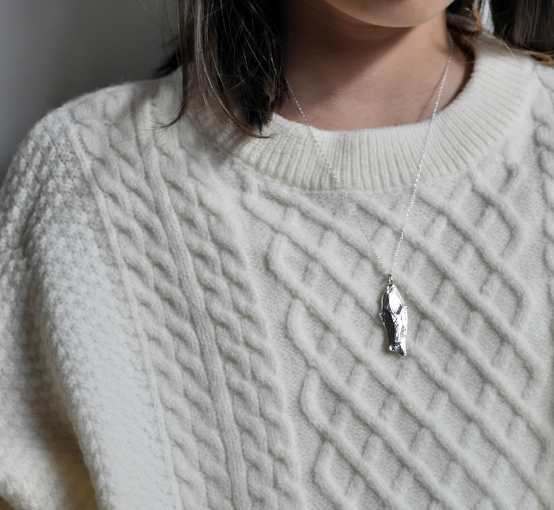 May include: A silver necklace with a unique, textured pendant shaped like a jagged rock. The necklace is worn by a person wearing a white cable knit sweater.