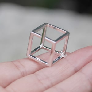 Può includere: Un anello in argento a forma di cubo geometrico con un centro vuoto. L'anello è realizzato con un filo sottile ed è progettato per assomigliare a un cubo tridimensionale.