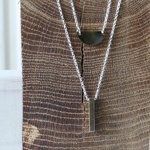 May include: A silver chain necklace with two pendants. The top pendant is a half-circle shape and the bottom pendant is a long, thin rectangle.