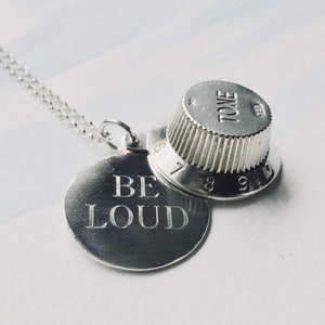 Puede incluir: Collar de plata con un colgante redondo que dice "BE LOUD" y un colgante de perilla de guitarra de plata con la palabra "TONE" en él.