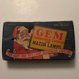 May include: A vintage box for Christmas lights featuring a Santa Claus illustration holding a sign that reads "GEM CHRISTMAS LIGHTS with MAZDA LAMPS". The box is blue with red and gold accents. The text "FOR SAFE AND BRILLIANT DECORATIVE LIGHTING" is printed on a white ribbon.