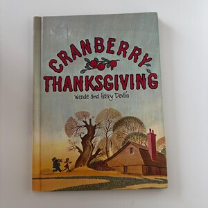May include: A hardcover book titled "Cranberry Thanksgiving" by Wende and Harry Devlin. The cover features red text and illustrations of cranberries, a house, and two figures running. The background is a light blue and green.