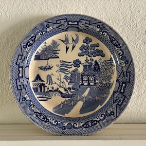 May include: A blue and white ceramic plate with a willow pattern design. The plate features a central image of a pagoda, trees, and a river with a boat. The border of the plate is decorated with a repeating pattern of stylized flowers and leaves.