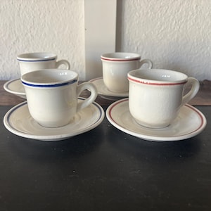 May include: Set of four off-white ceramic espresso cups and saucers. Each cup and saucer has a colored band around the rim: blue, red, or a combination of both. The cups have small handles.