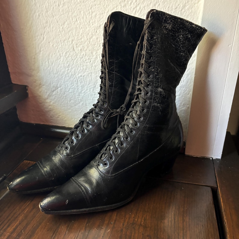 May include: A pair of black, vintage lace-up boots. The boots have pointed toes and a stacked heel. The leather appears worn, with visible creases and wear. The laces are tightly laced up the front of the boots.