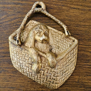 May include: A brass door knocker shaped like a dog in a basket. The dog is sitting up with its head tilted to the side and its tongue sticking out. The basket is woven with a pattern of squares and diamonds.