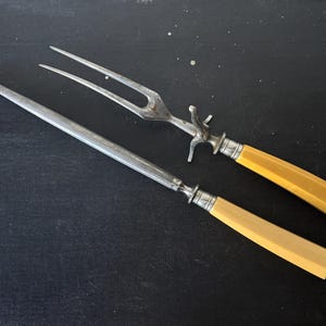 May include: Antique carving set with a fork and a sharpening steel. The set features silver-toned metal blades and handles with a light yellow, rectangular design. The fork has three tines and a decorative bolster.