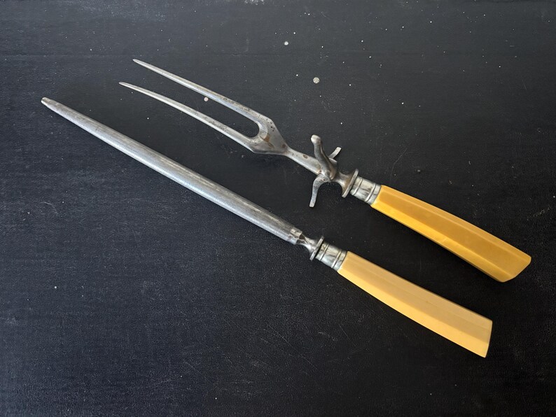 May include: Antique carving set with a fork and a sharpening steel. The set features silver-toned metal blades and handles with a light yellow, rectangular design. The fork has three tines and a decorative bolster.