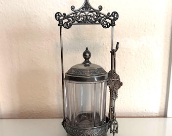 May include: An antique silver-toned sugar bowl with a clear, ribbed glass container. The bowl has a decorative lid and sits in a detailed metal frame with a handle and tongs. The design features ornate scrollwork.