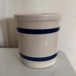 May include: A cylindrical stoneware crock with a cream-colored base and two navy blue bands. The vintage kitchenware has a glossy finish and a simple, classic design. This piece is ideal for rustic or farmhouse decor.
