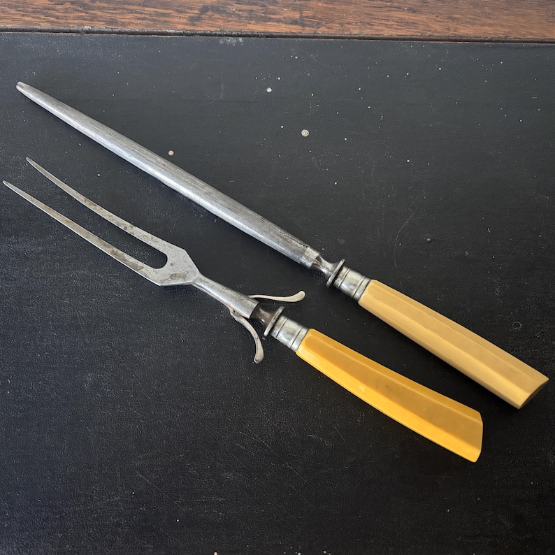 May include: A carving fork and a sharpening steel are displayed. The fork has two tines and a light-colored handle. The sharpening steel has a long, metal rod and a matching handle. Both have silver-colored metal accents.