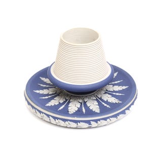 May include: A blue and white ceramic decorative piece. The base is a round plate with a detailed leaf pattern. A white, ribbed, conical cup sits atop the plate. The piece is likely a decorative item or a small container.
