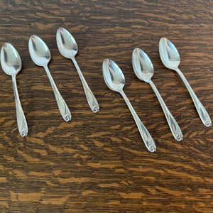 May include: Six silver teaspoons with ornate floral designs on the handles. The spoons are arranged in a row on a wooden surface.