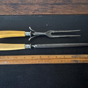 May include: Antique carving set with a fork and a sharpening steel, both with cream-colored handles and silver accents. A wooden ruler is also present. The set is displayed on a dark surface.