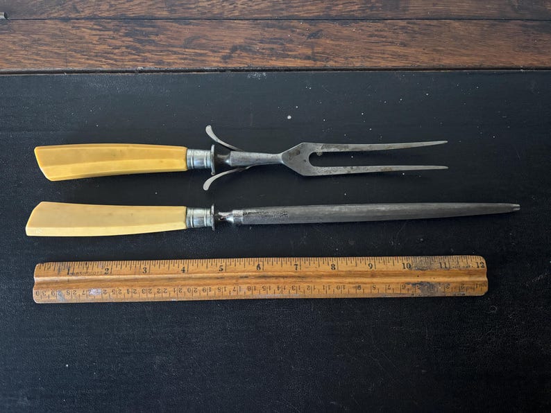 May include: Antique carving set with a fork and a sharpening steel, both with cream-colored handles and silver accents. A wooden ruler is also present. The set is displayed on a dark surface.