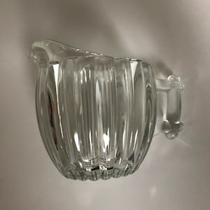 May include: A clear glass creamer with a ribbed design and a handle. The creamer is perfect for serving cream or milk.