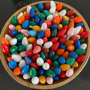May include: A bowl filled with a variety of colorful light bulbs. The bulbs are in various shapes and sizes, including round, teardrop, and elongated. The colors include red, orange, yellow, green, blue, purple, and white.