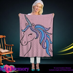 Puede incluir: Una manta de ganchillo rectangular con fondo rosa y un gráfico grande de una cabeza de unicornio. El unicornio tiene una melena azul y morada, un cuerno azul y ojos cerrados. El texto "Digital mockup shown" está impreso en la esquina inferior derecha.